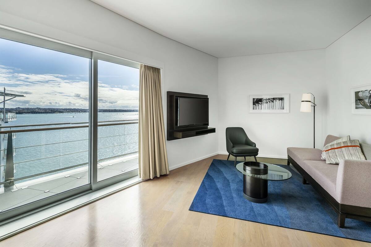 A modern hotel room with a large window overlooking a body of water, a flat-screen TV mounted on the wall, a sofa, a chair, and a small coffee table.
