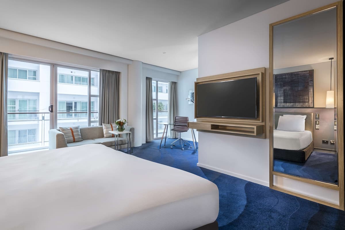 A hotel room with a large bed, a TV mounted on the wall, a small table with a chair, and a balcony with curtains.
