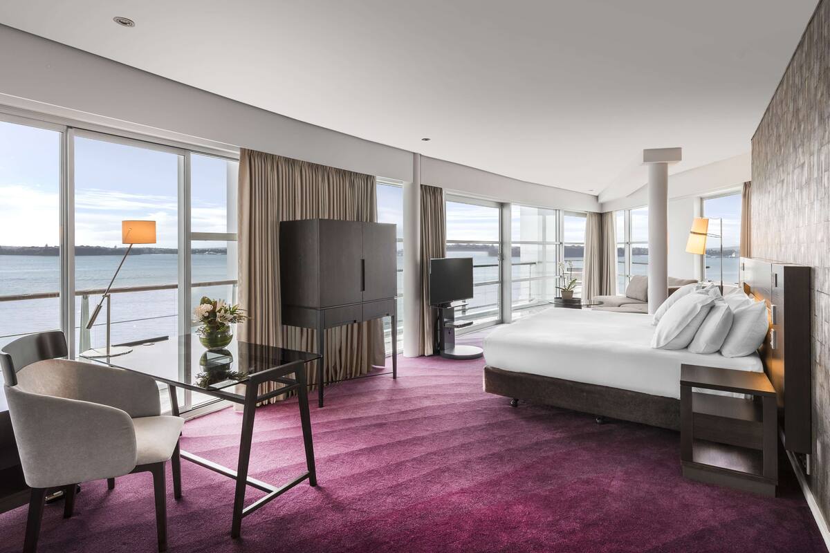 A modern hotel room with a large bed, a TV, a glass table, and a view of the water.