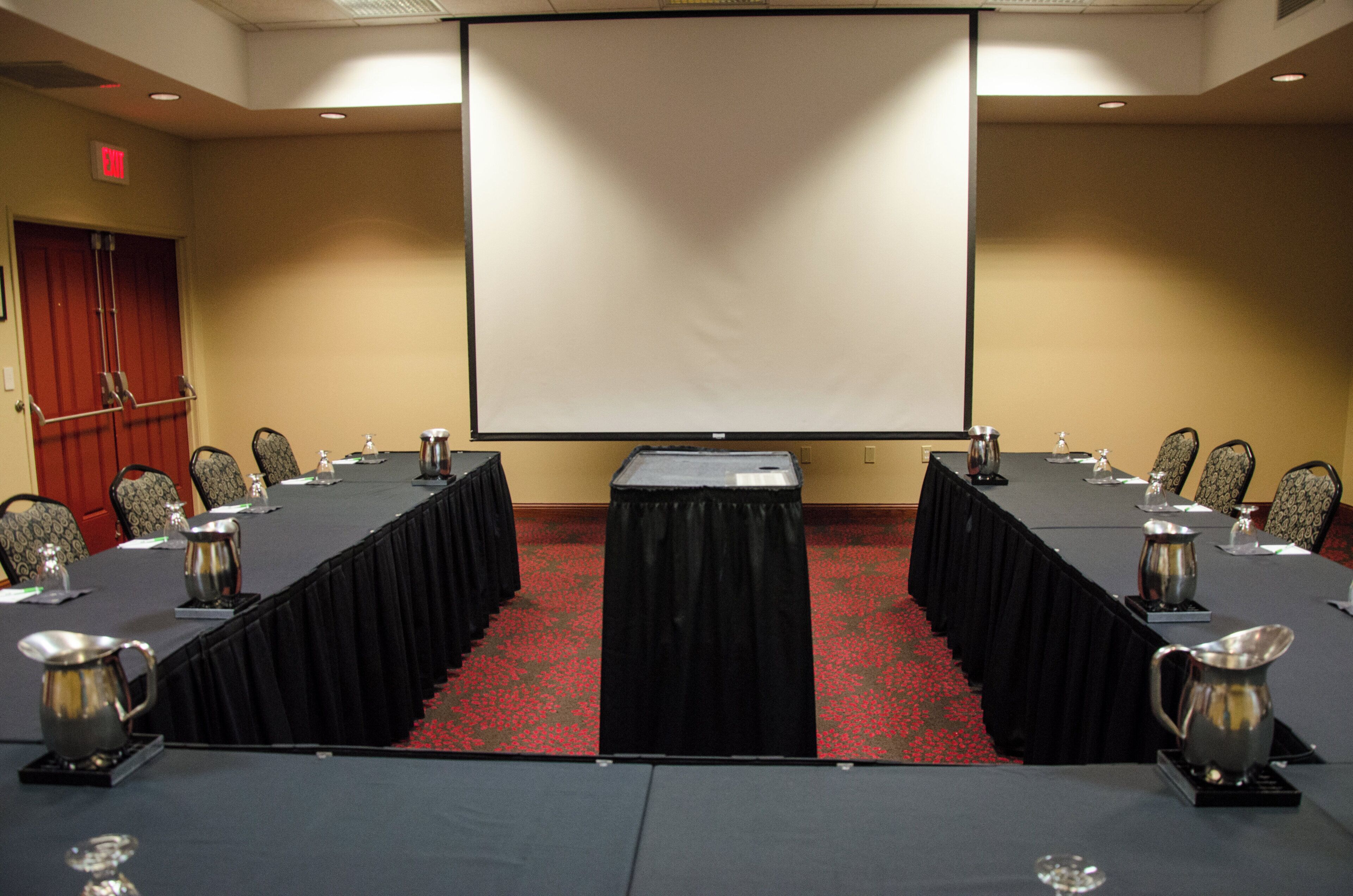 A conference room with a long table, chairs, a projector screen, and a red carpeted floor.
