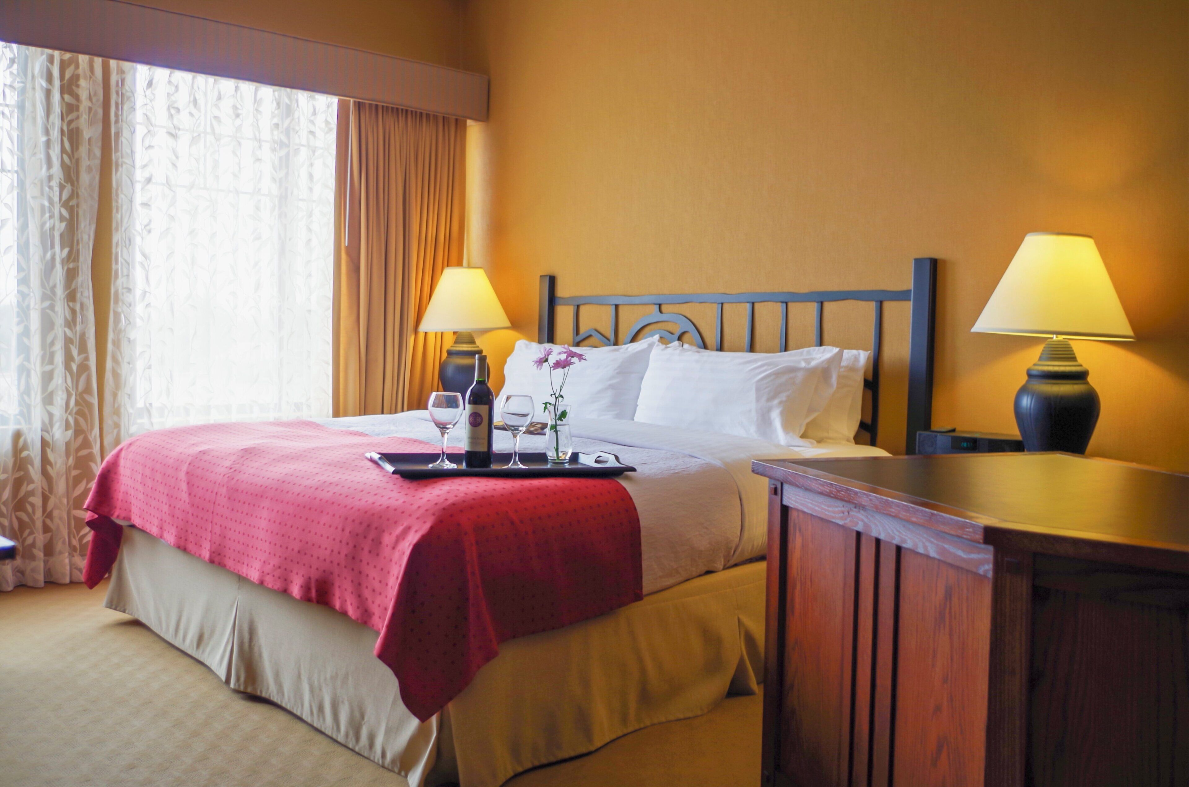 A hotel room with a bed, two bedside lamps, a nightstand, a tray with a bottle and glasses, and a window with curtains.