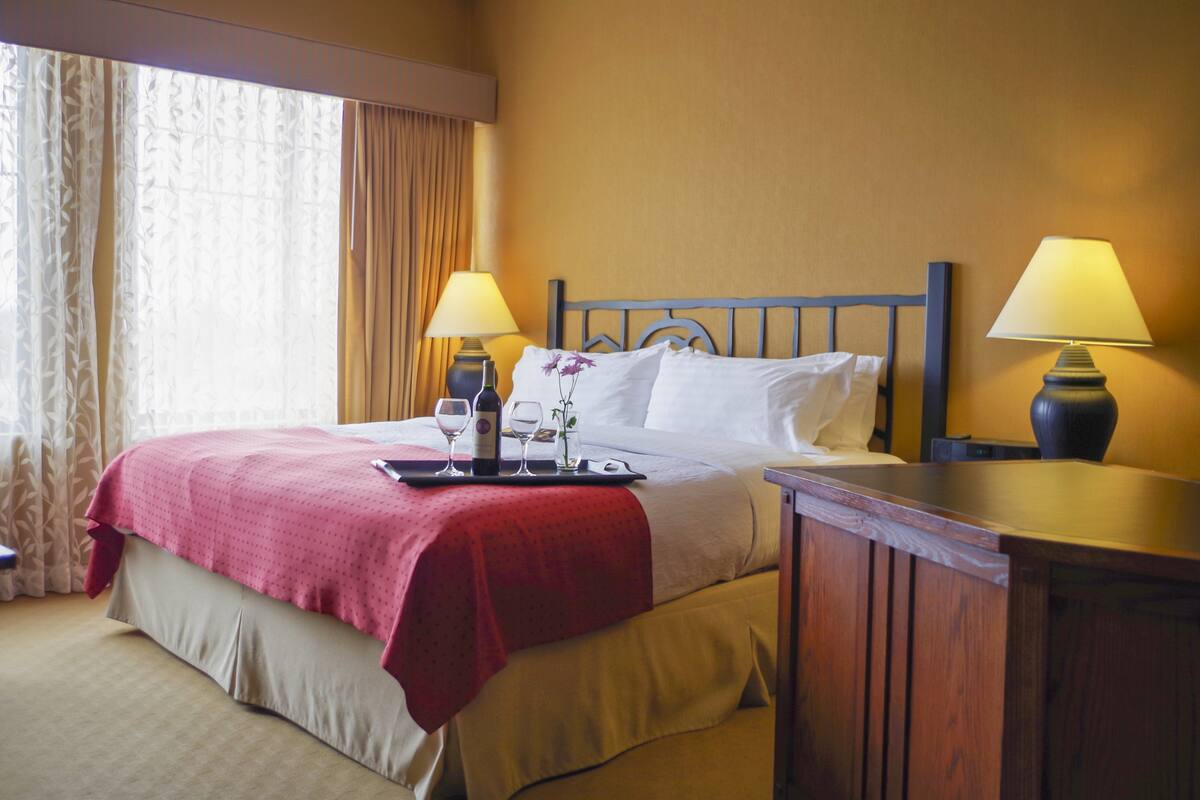 Una habitación de hotel con una cama, dos lámparas de noche, una bandeja con vasos y una botella, y una mesita de noche de madera.