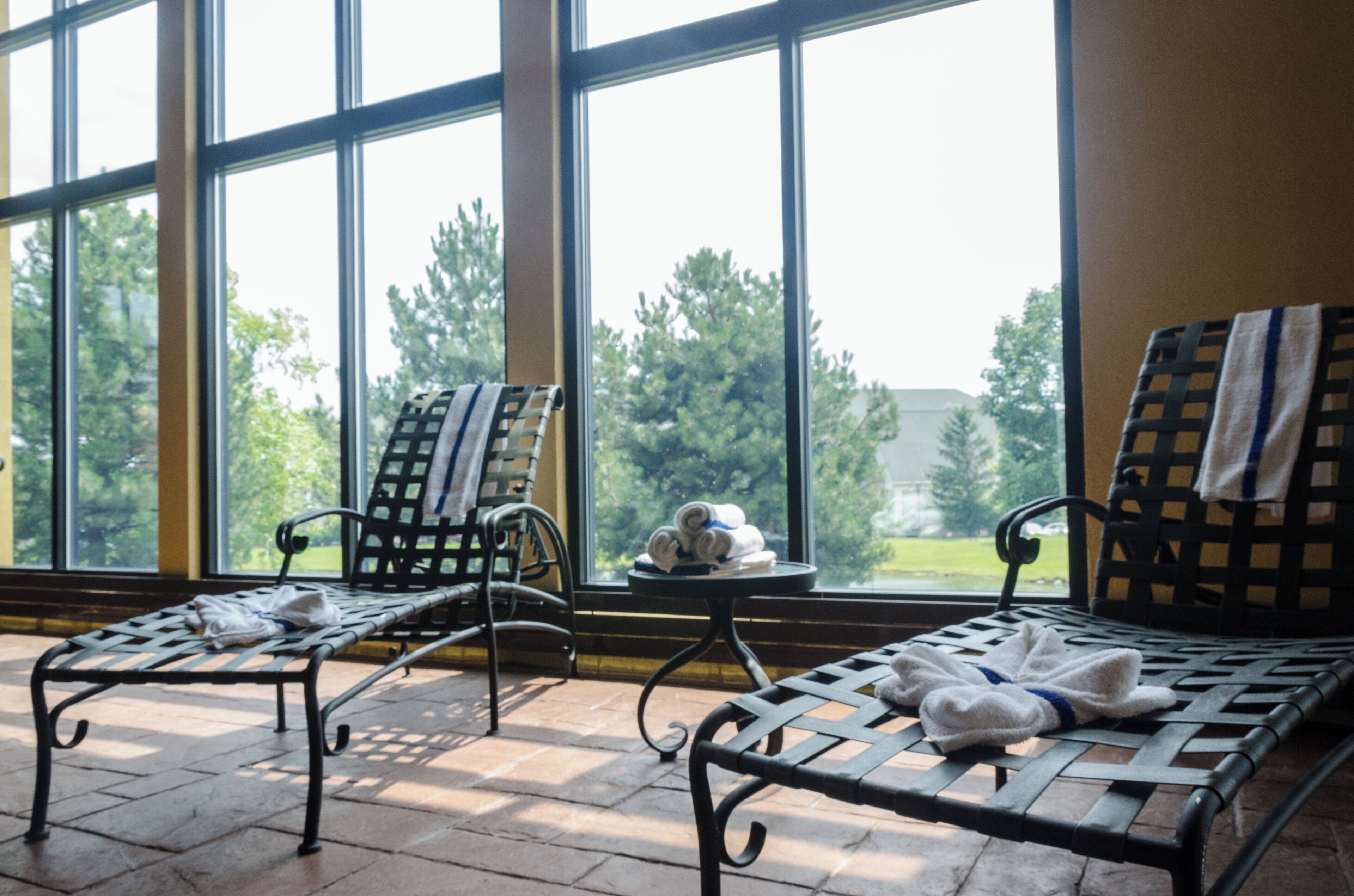A room with two metal-framed lounge chairs, one with a towel on it, and a small table with a towel and a rolled-up item.