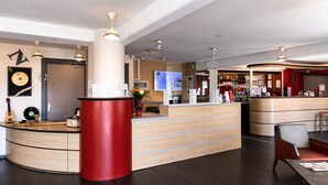 A modern hotel lobby with a curved reception desk, a red accent column, and a bar area in the background.