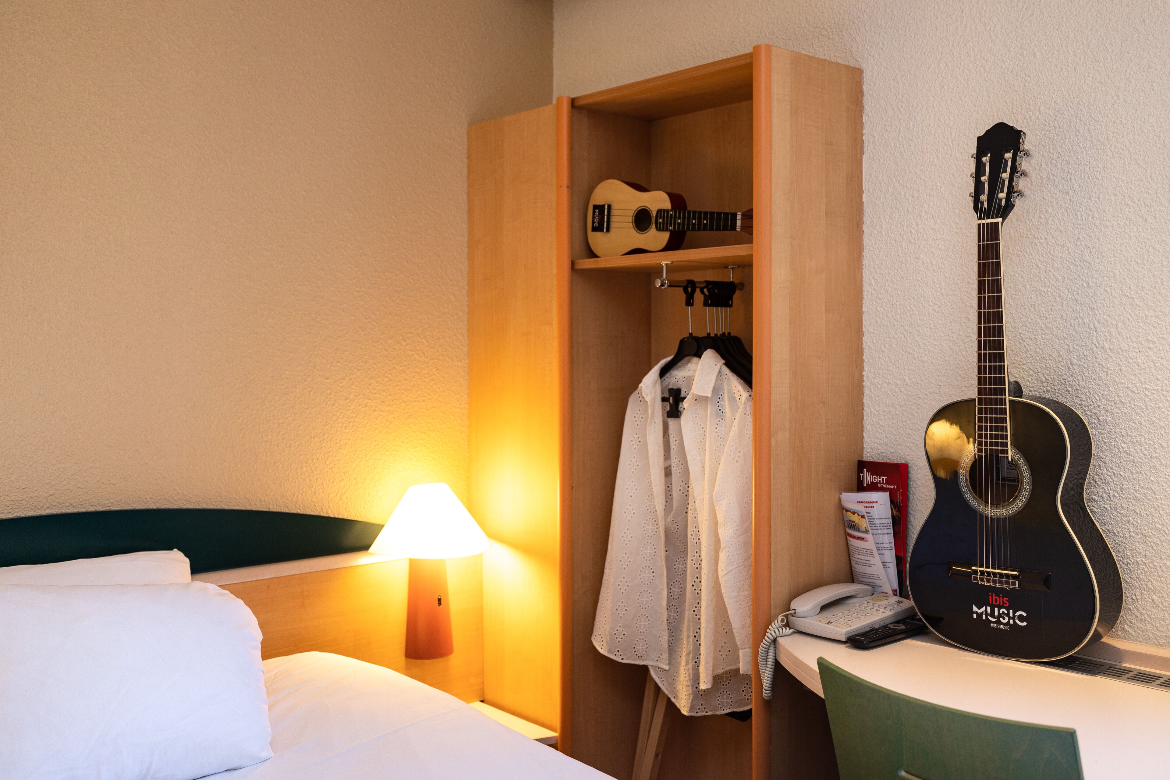 A hotel room with a bed, a wooden wardrobe, a guitar, a lamp, and a telephone.
