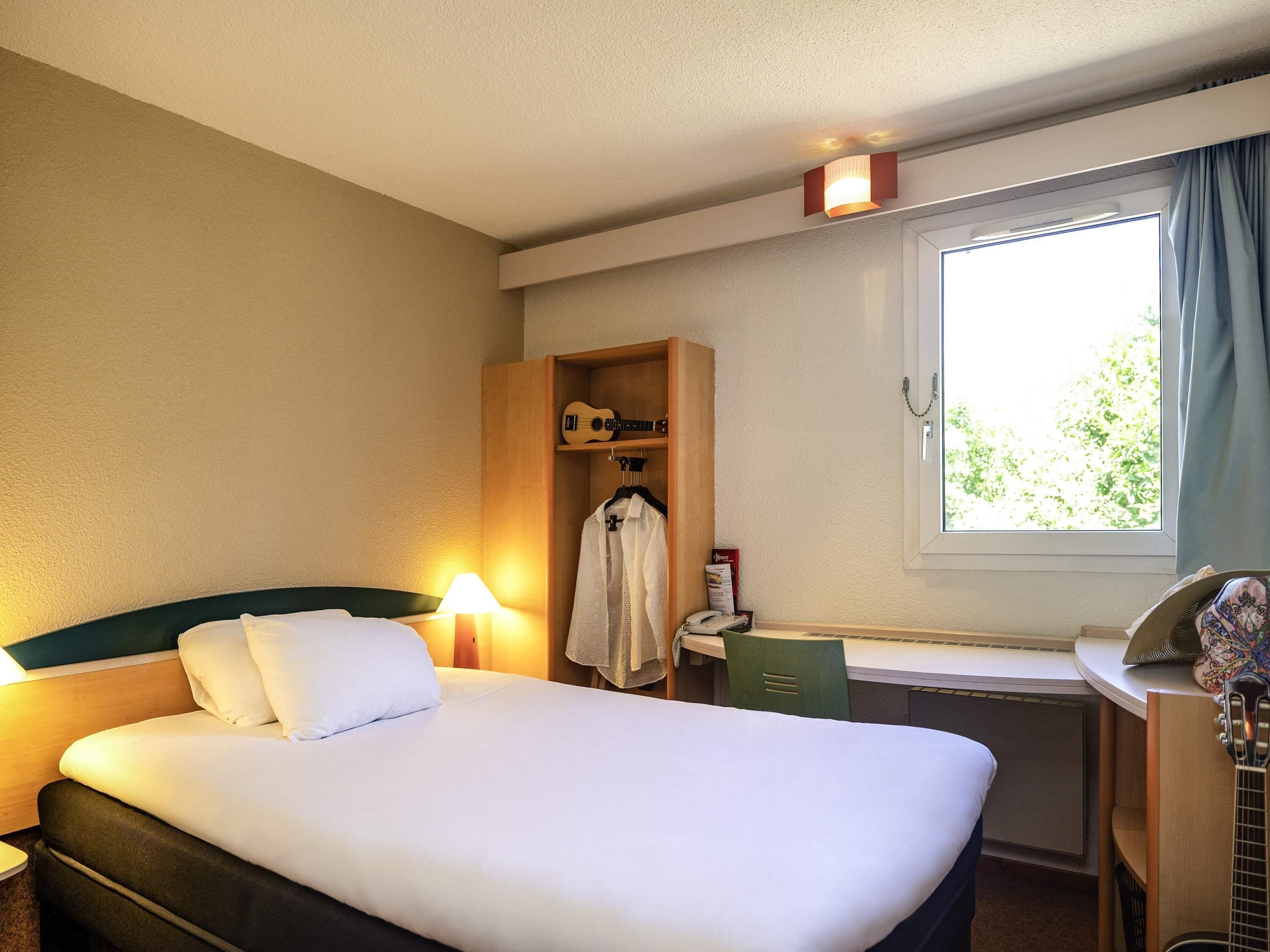 A hotel room with a bed, a desk, a guitar, and a window with curtains.
