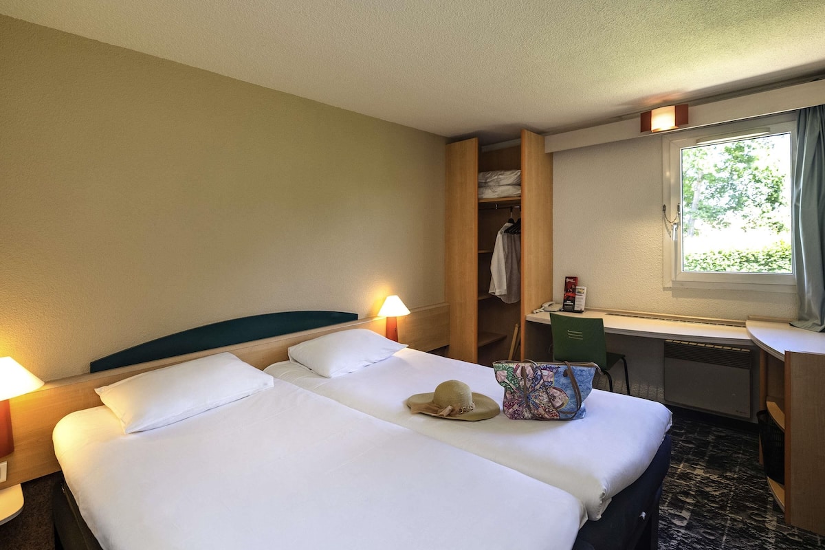 A hotel room with two beds, a desk, a chair, a window with a view of greenery, and a closet with clothes.