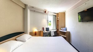 A hotel room with a bed, a desk with a computer, a TV, and a window with curtains.