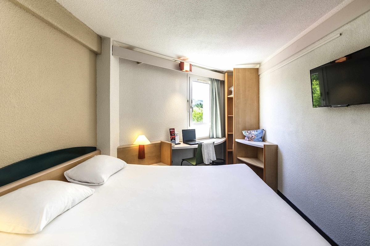 A hotel room with a bed, a desk with a computer, a TV, and a window with curtains.
