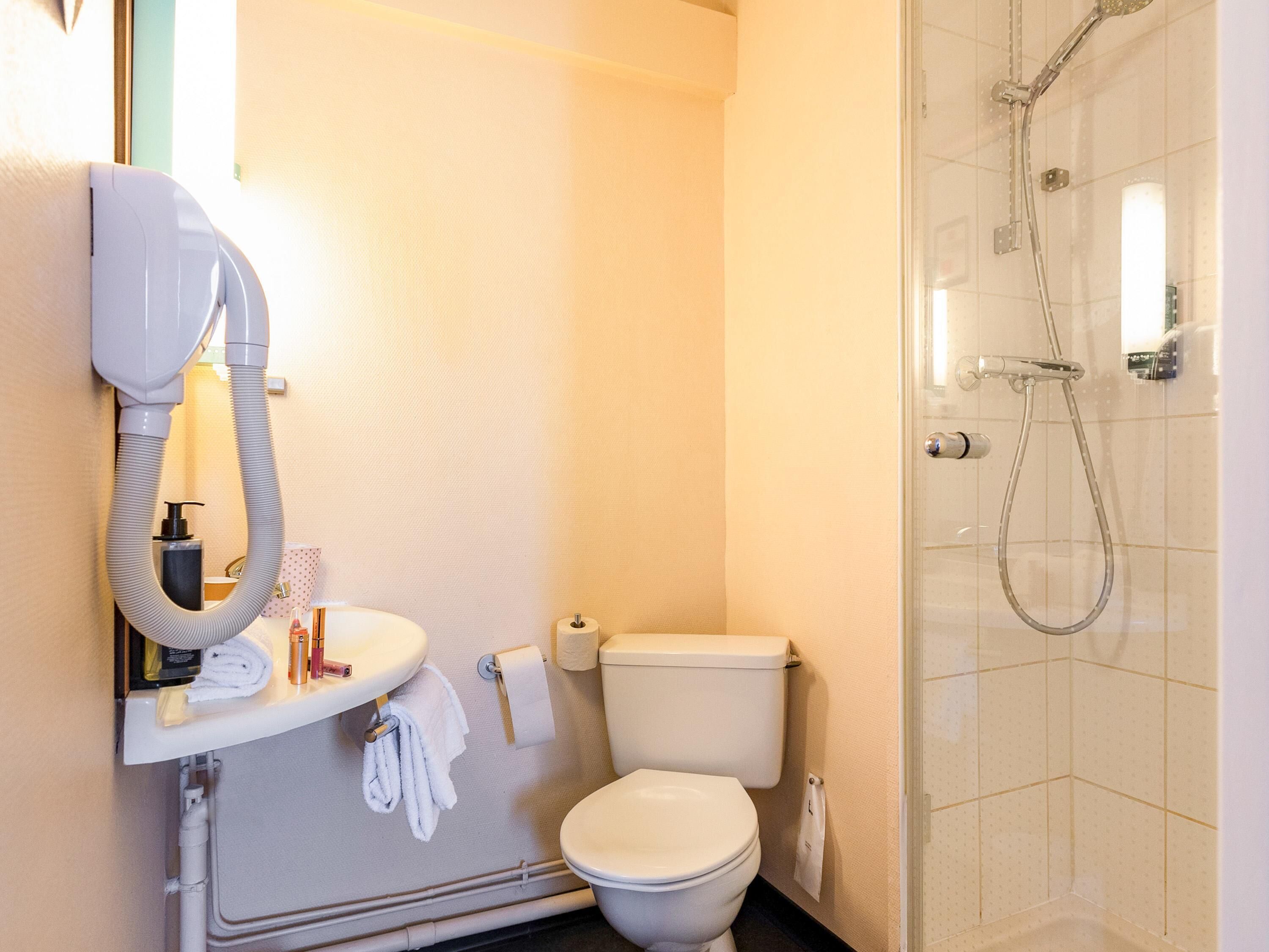 Une salle de bain avec des toilettes blanches, un lavabo équipé d’un sèche-cheveux et un espace douche.