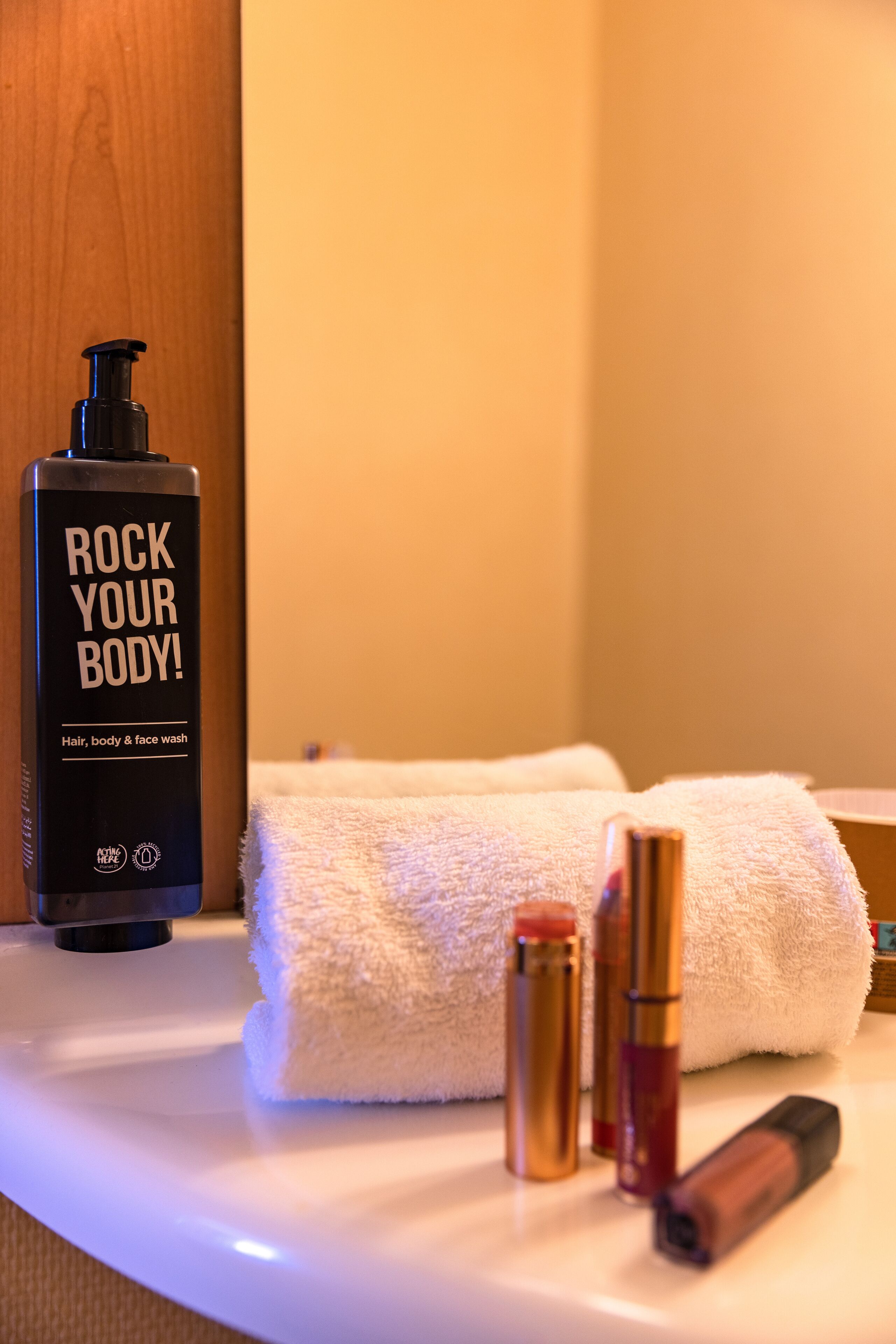 Ein Badezimmer-Waschtisch mit einer Flasche mit dem Aufdruck "ROCK YOUR BODY!", einem weißen Handtuch und mehreren Kosmetikbehältern.