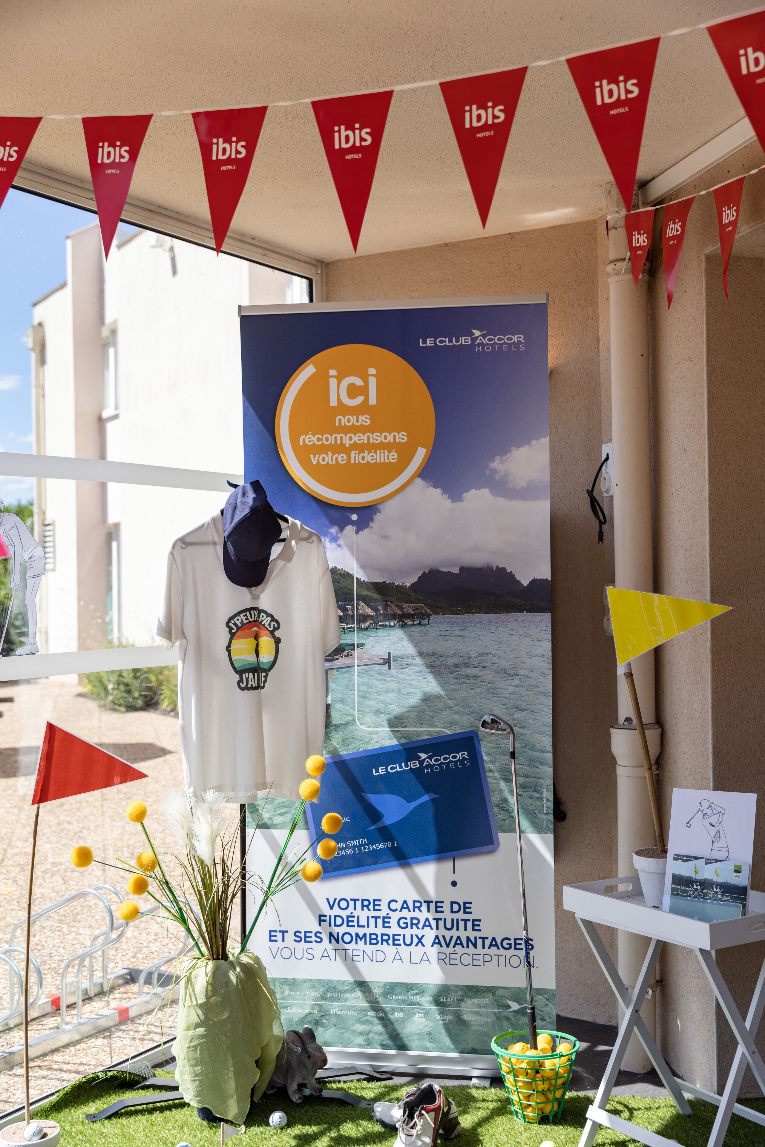 Entrada de hotel con un cartel para Le Club Accor Hotels, un letrero de bienvenida y una exhibición de servicios que incluye un mapa, un sombrero y una pequeña estatua.