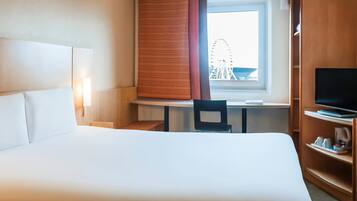 A hotel room with a large bed, a desk with a chair, a television on a stand, and a window with a view of a building.
