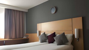 A hotel room with a large bed, a bedside table, a clock on the wall, and a window with curtains.