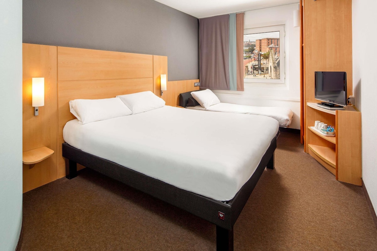 A hotel room with a large bed, bedside tables, a TV on a stand, and a view of the city through the window.