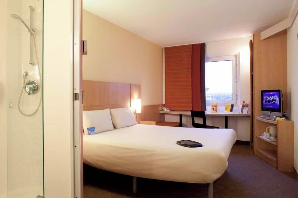 A hotel room with a bed, a desk with a chair, a television, and a shower area.