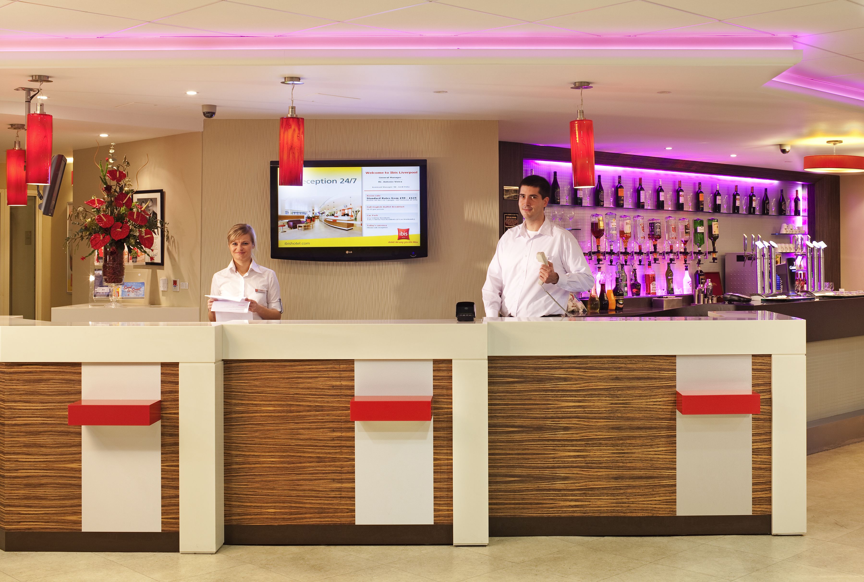 Ein Hotel-Empfangsbereich mit einer Rezeption, einem Bildschirm, der Informationen zeigt, und einer Bar mit einer Auswahl an Flaschen.
