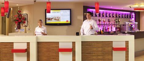 A hotel reception area with a reception desk, a television screen displaying information, and a bar with various bottles.