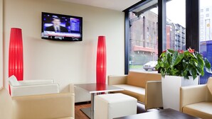 A modern hotel lobby with a TV mounted on the wall, red accent chairs, a white sofa, a small table, and a potted plant.