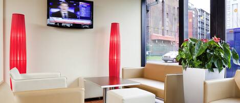 A modern hotel lobby with a TV mounted on the wall, red accent chairs, a white sofa, a small table, and a potted plant.
