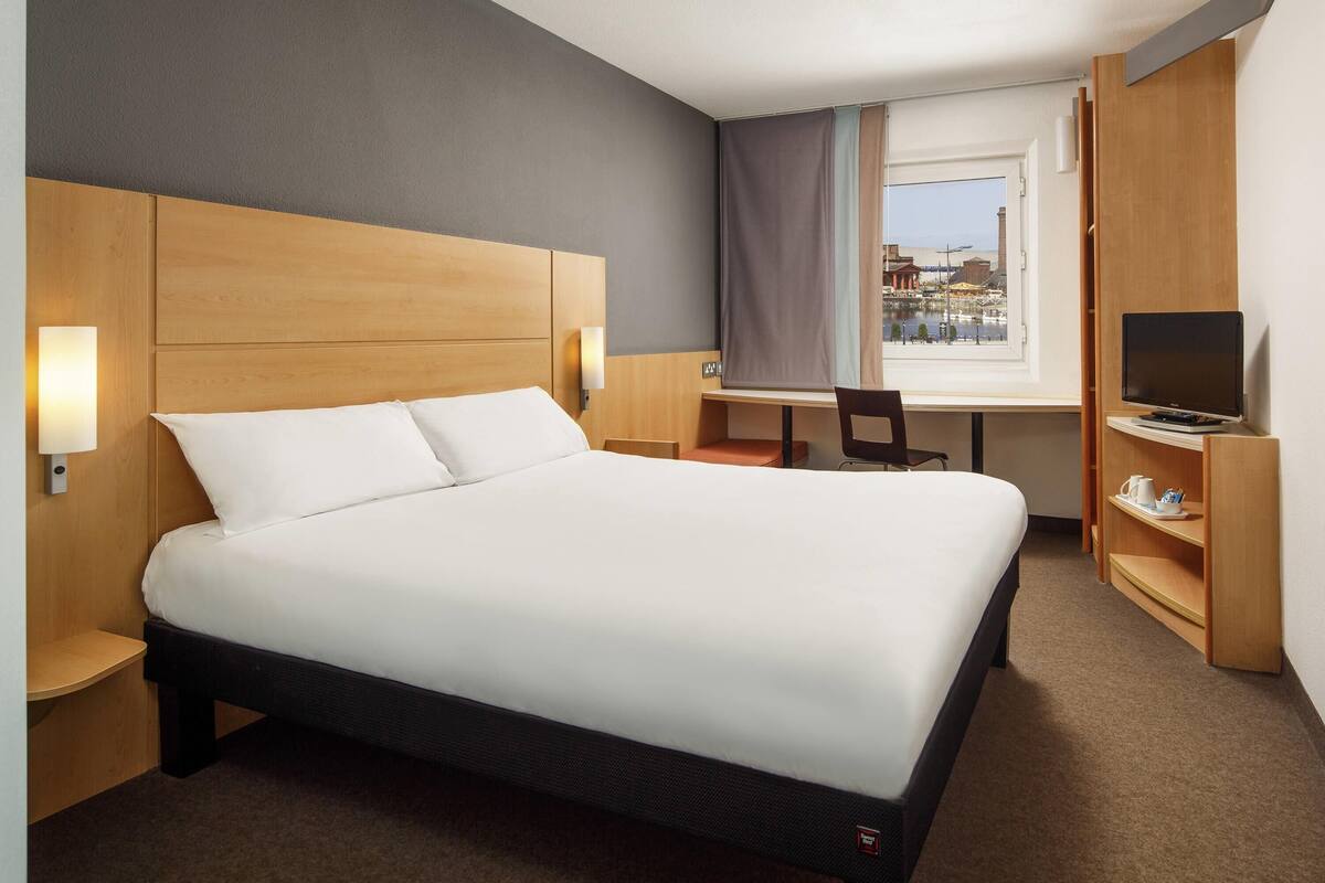 A hotel room with a large bed, a desk, a chair, a TV, and a view of the city.