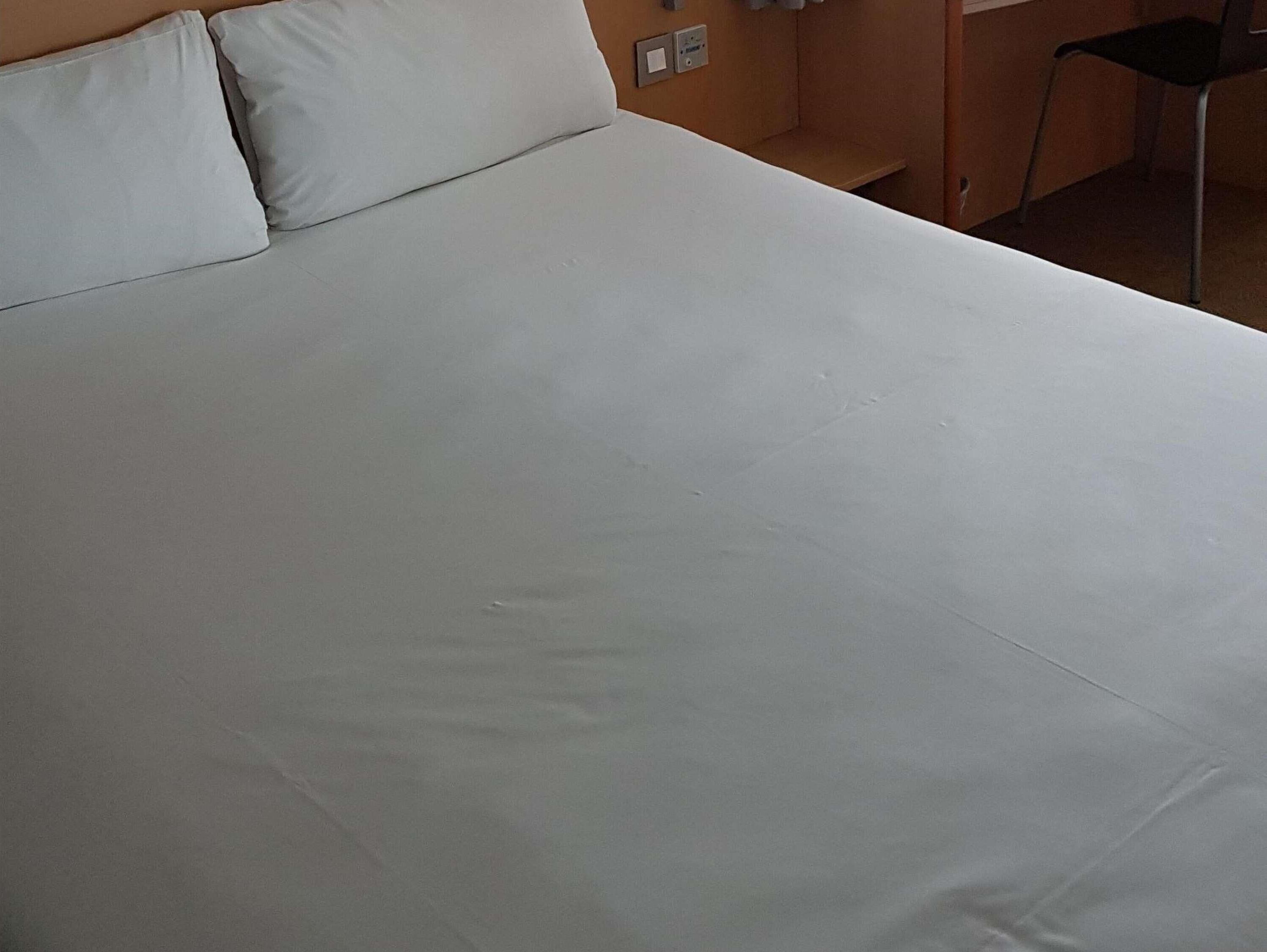 A bed with white linens, a wooden headboard, and a wall-mounted light switch.