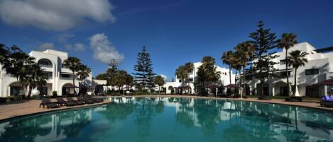 A pool surrounded by lounge chairs and palm trees, with a clear blue sky above.
