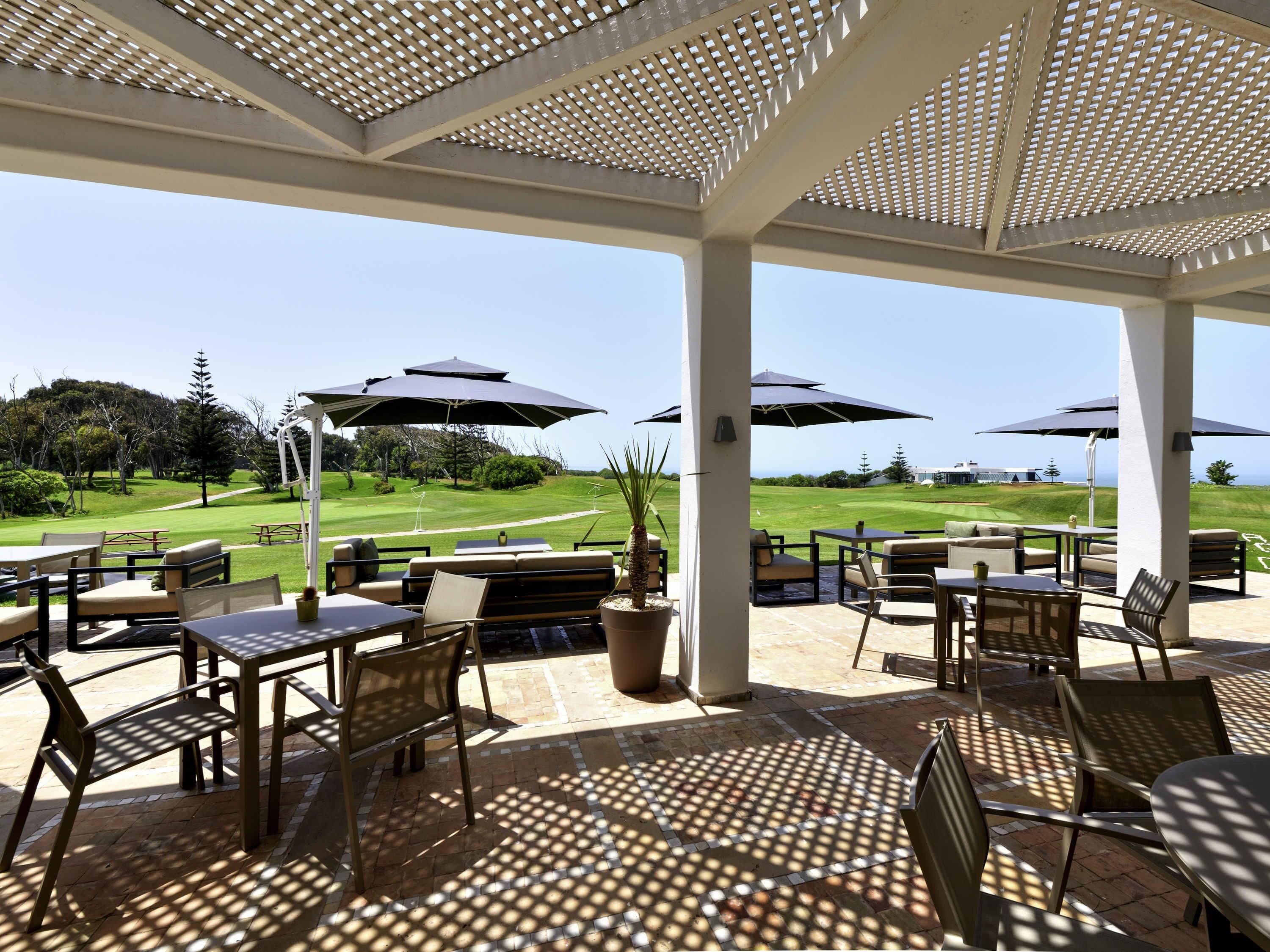 A covered outdoor seating area with tables and chairs, umbrellas, and a view of a golf course.