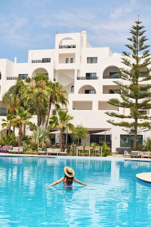 Una piscina con una persona nadando, rodeada de palmeras y un edificio blanco con balcones.