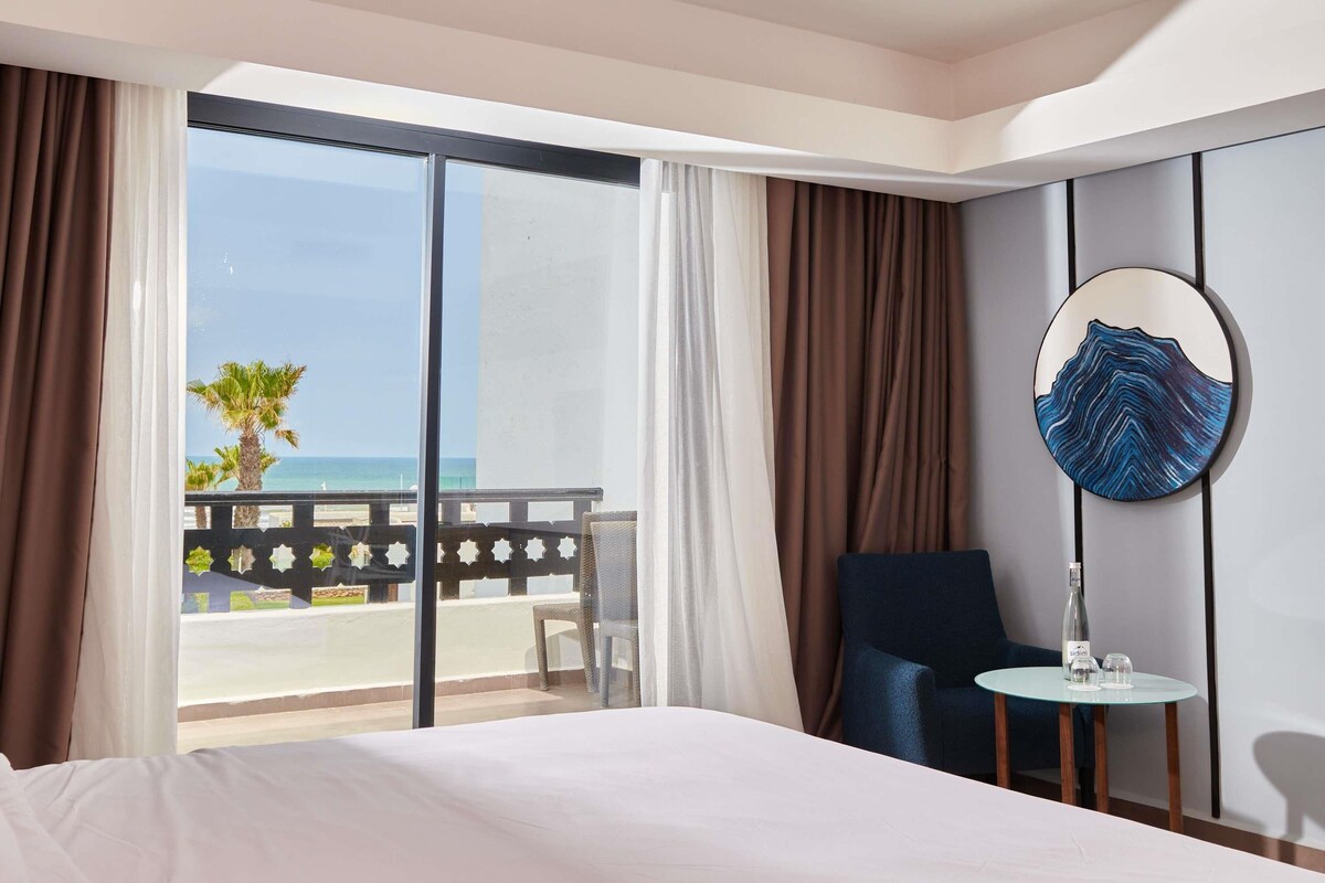 Una habitación de hotel con cama, una silla, una mesita con una botella y vasos, un espejo redondo y un balcón con vistas al mar.