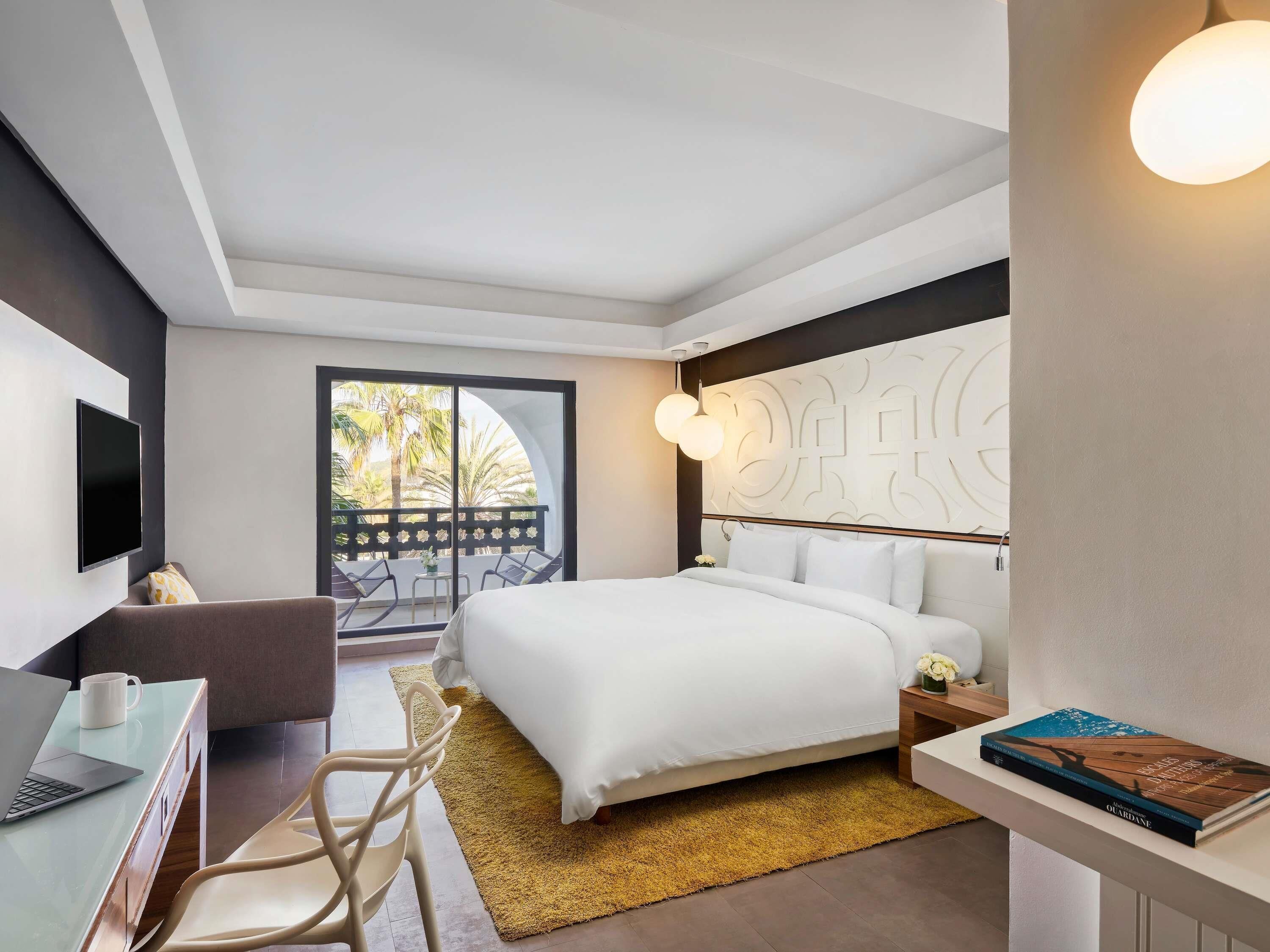 Ein modernes Hotelzimmer mit Bett, Schreibtisch mit Laptop, Stuhl, Nachttisch und einem Buch.