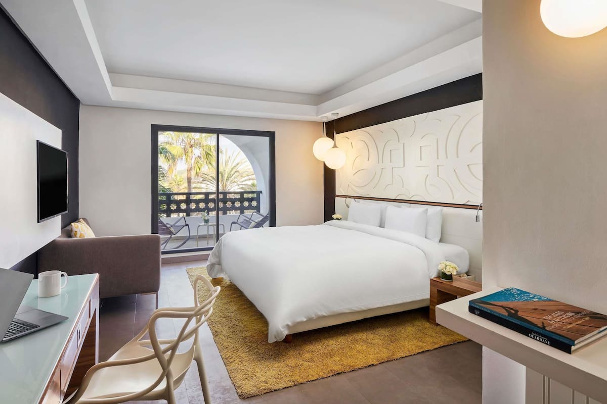 Een moderne hotelkamer met een bed, een bureau met een laptop, een stoel, een nachtkastje en een boek.