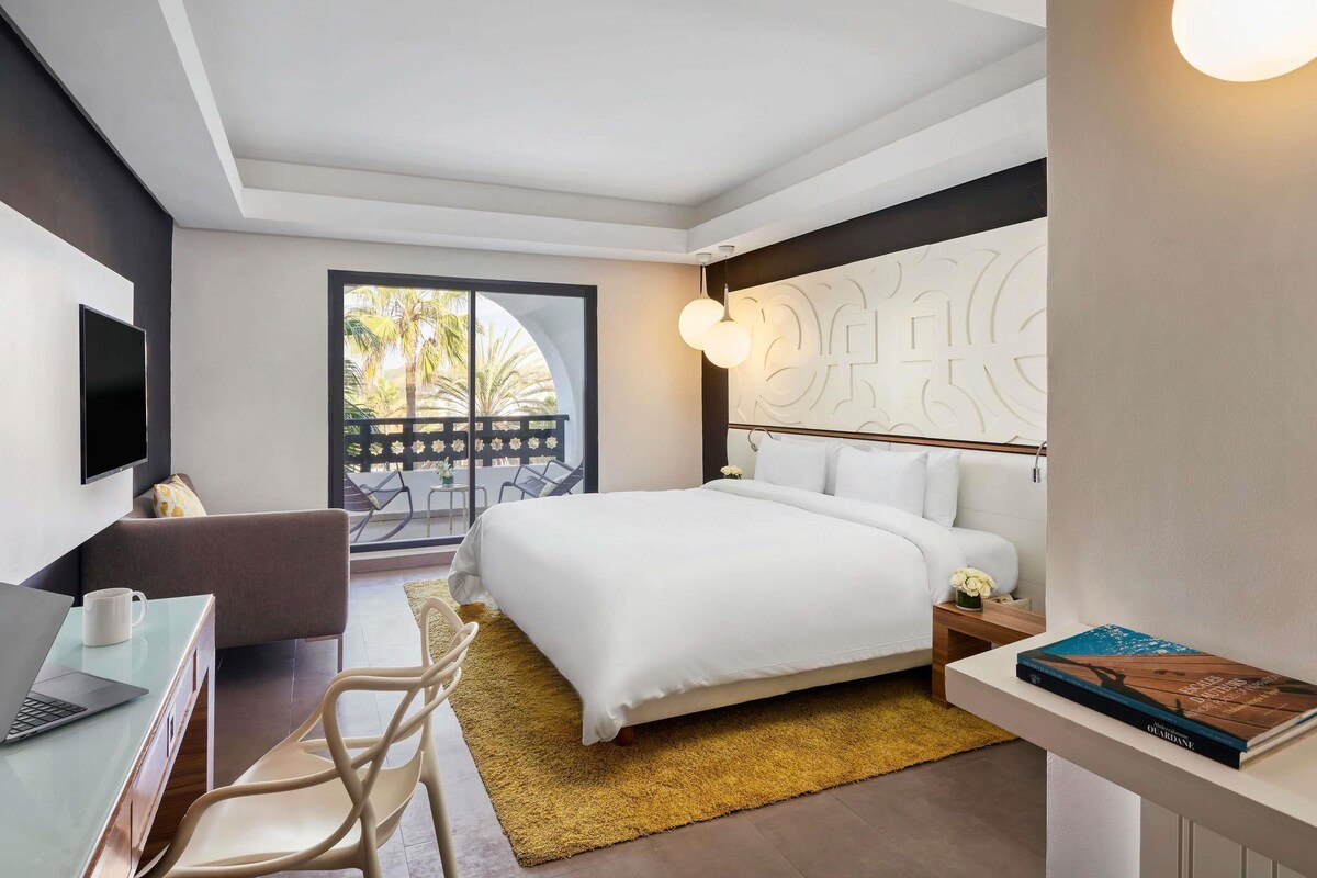 Una habitación de hotel moderna con una cama, un escritorio con una computadora portátil, una silla, una mesita de noche y un libro.