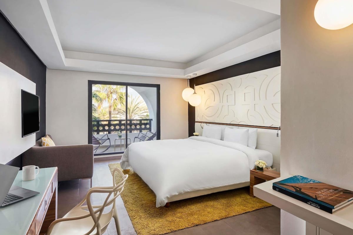 Ein modernes Hotelzimmer mit Bett, Schreibtisch mit Laptop, Stuhl, Fernseher und Balkon mit Blick auf Palmen.