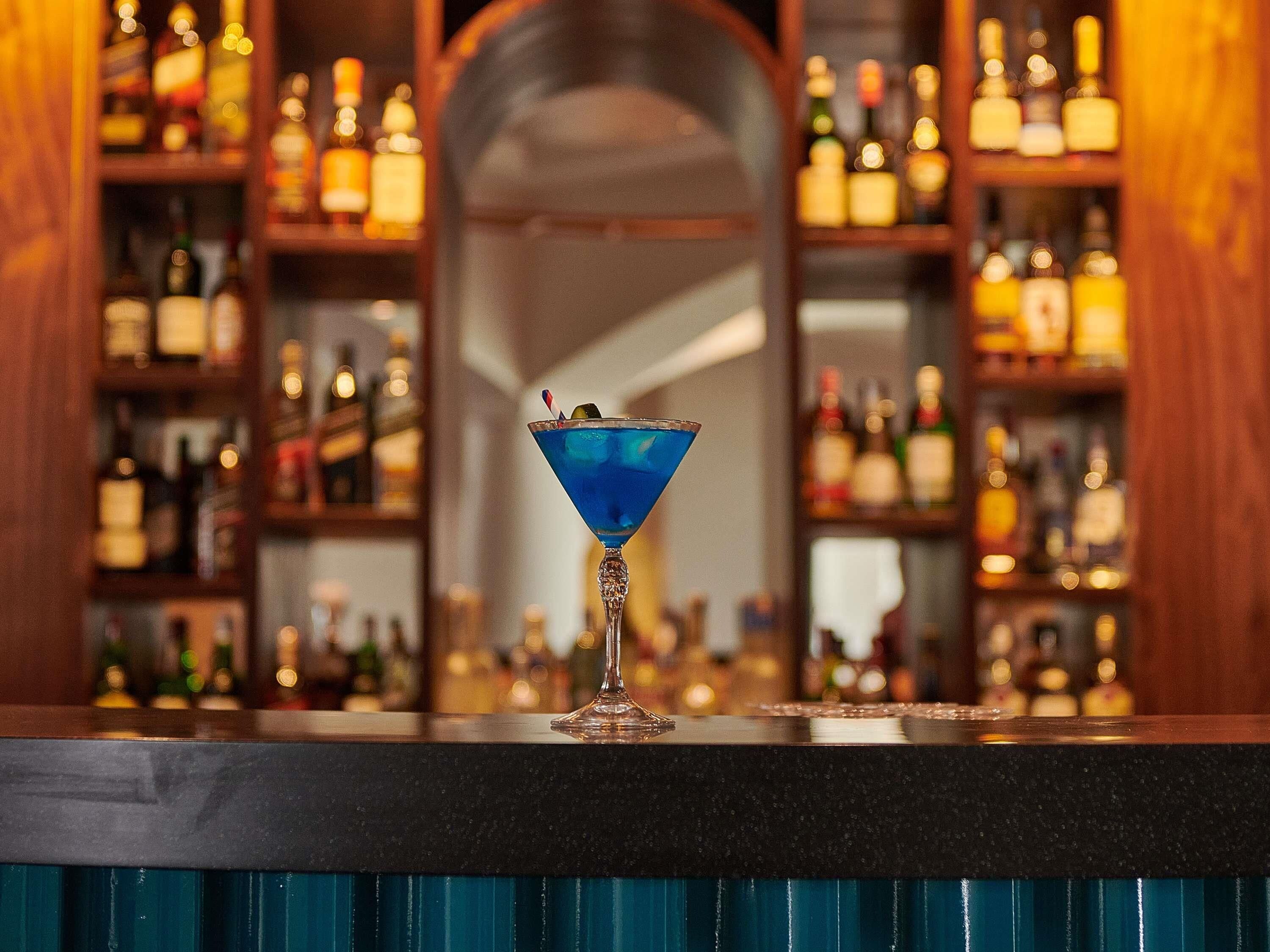 Zona de bar con un cóctel azul en una copa de martini, estantes llenos de botellas de licor y una barra de madera.