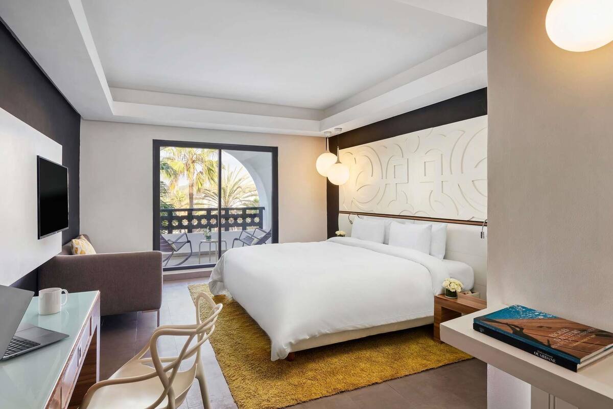 A modern hotel room with a bed, a desk with a laptop, a chair, a TV, and a balcony with a view of palm trees.