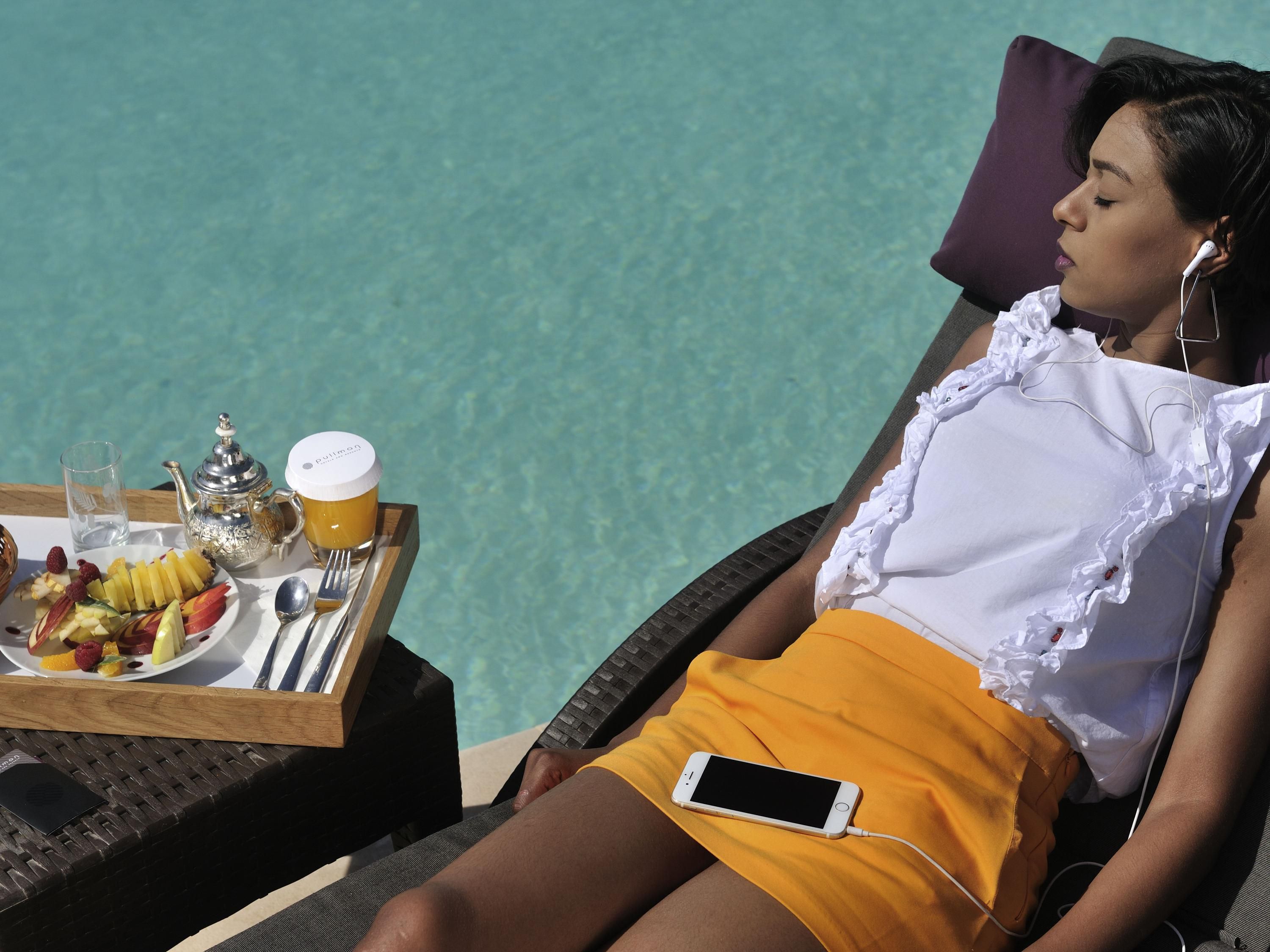 A person relaxing by a poolside with a tray of food, a teapot, and a glass of juice.