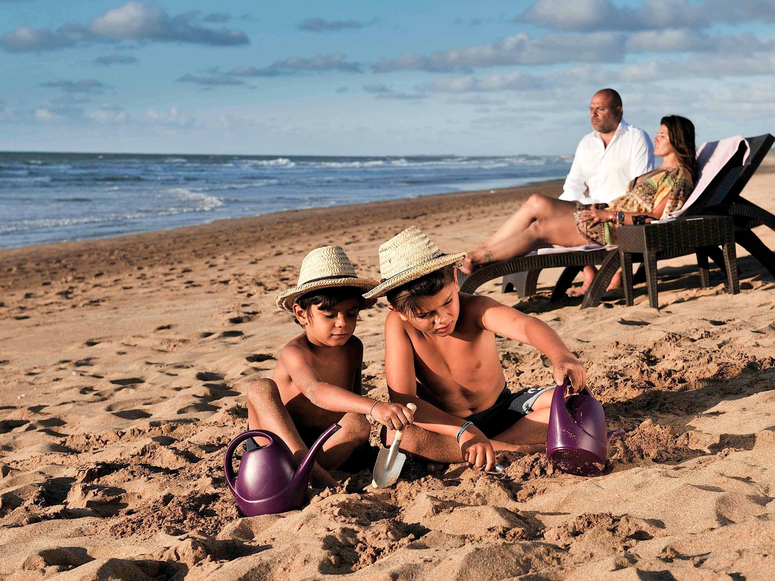 Une scène de plage où l’on voit des gens, dont des enfants qui jouent dans le sable, et un couple qui se détend sur des chaises longues.