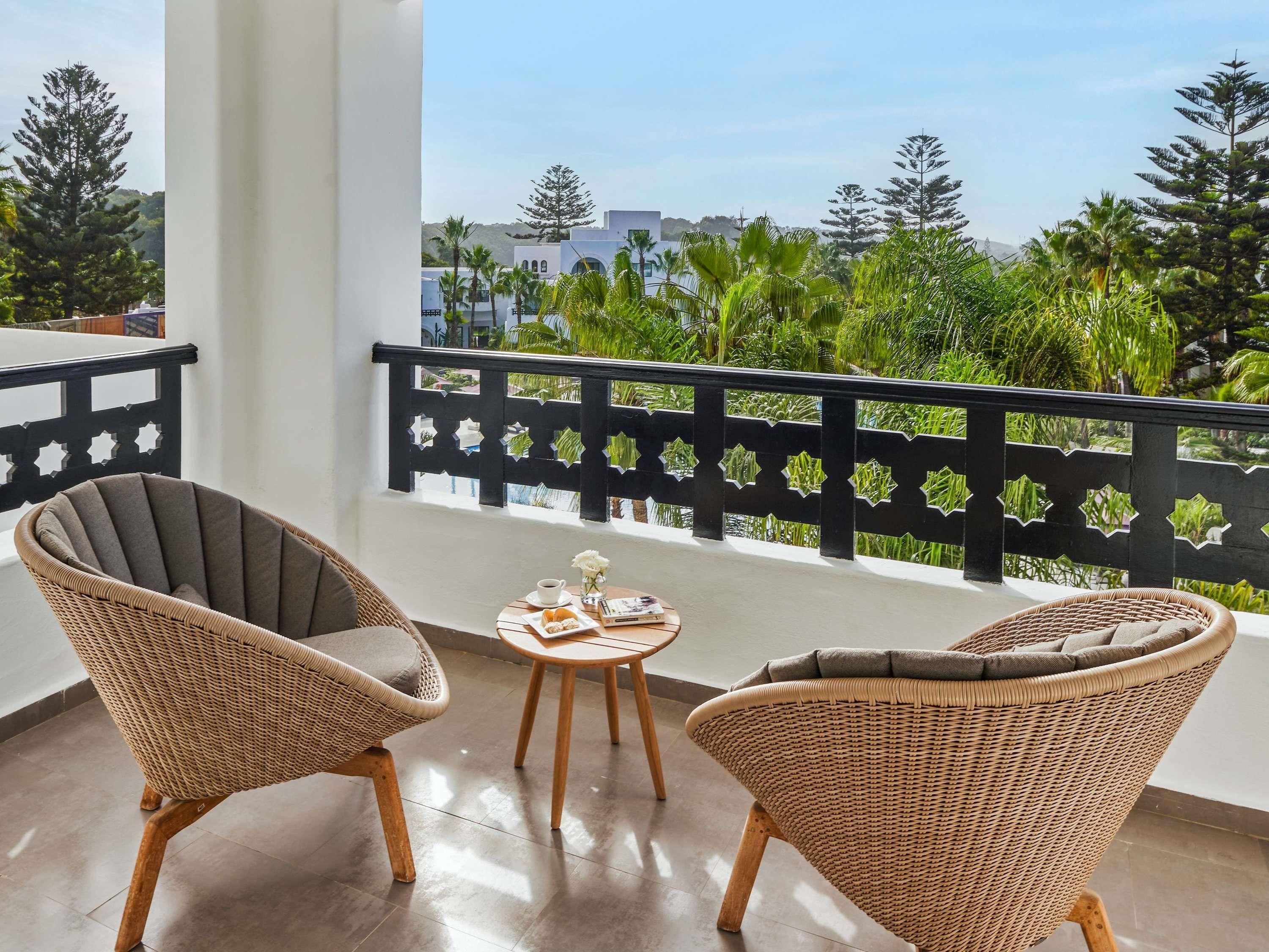 Ein Balkon mit Korbstühlen, einem kleinen Tisch mit Tasse und Gebäck, und Blick auf Grünflächen und Gebäude.
