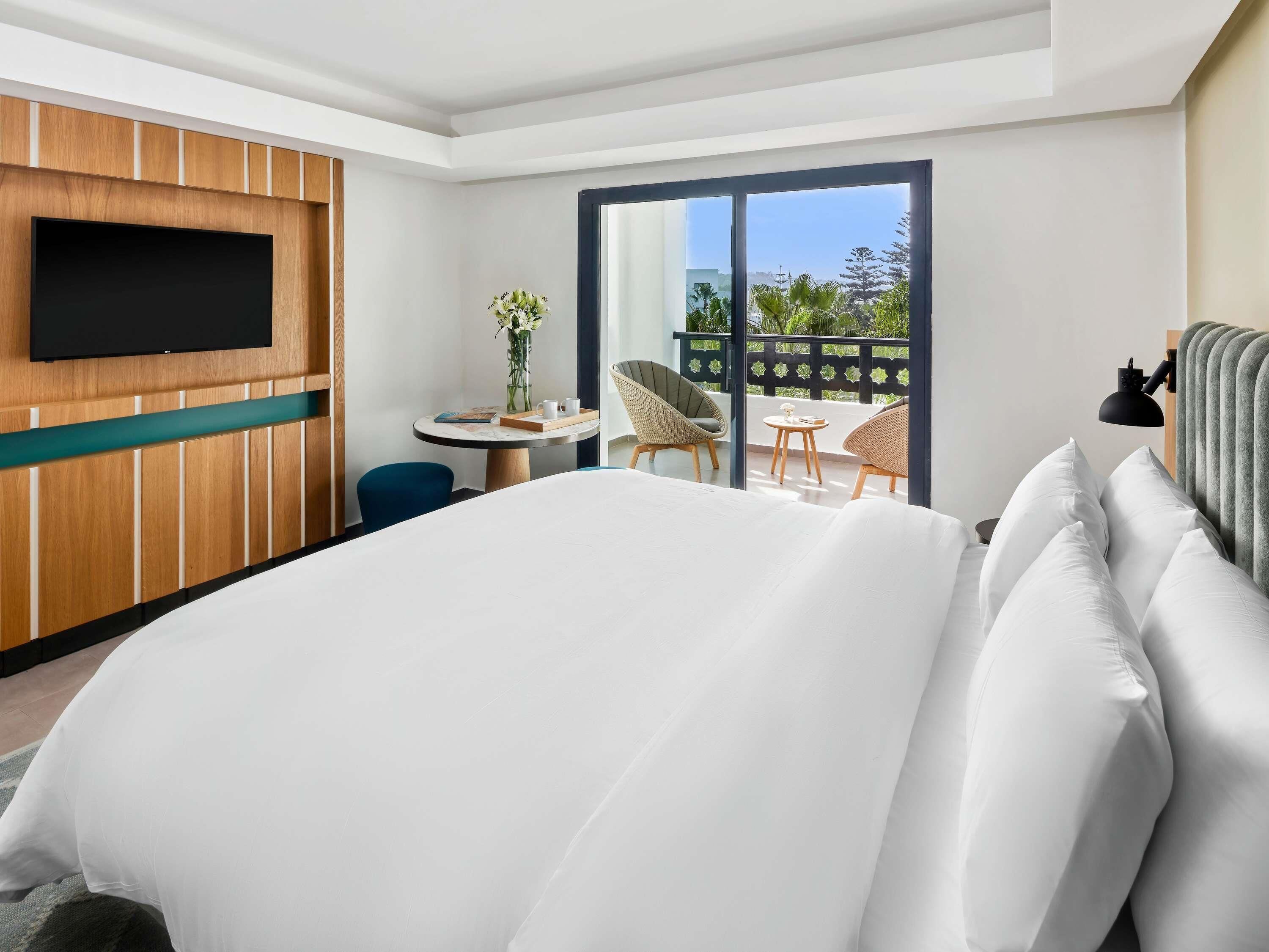 A hotel room with a large bed, a TV mounted on the wall, a small table with a vase of flowers, two wicker chairs, and a balcony with a view.