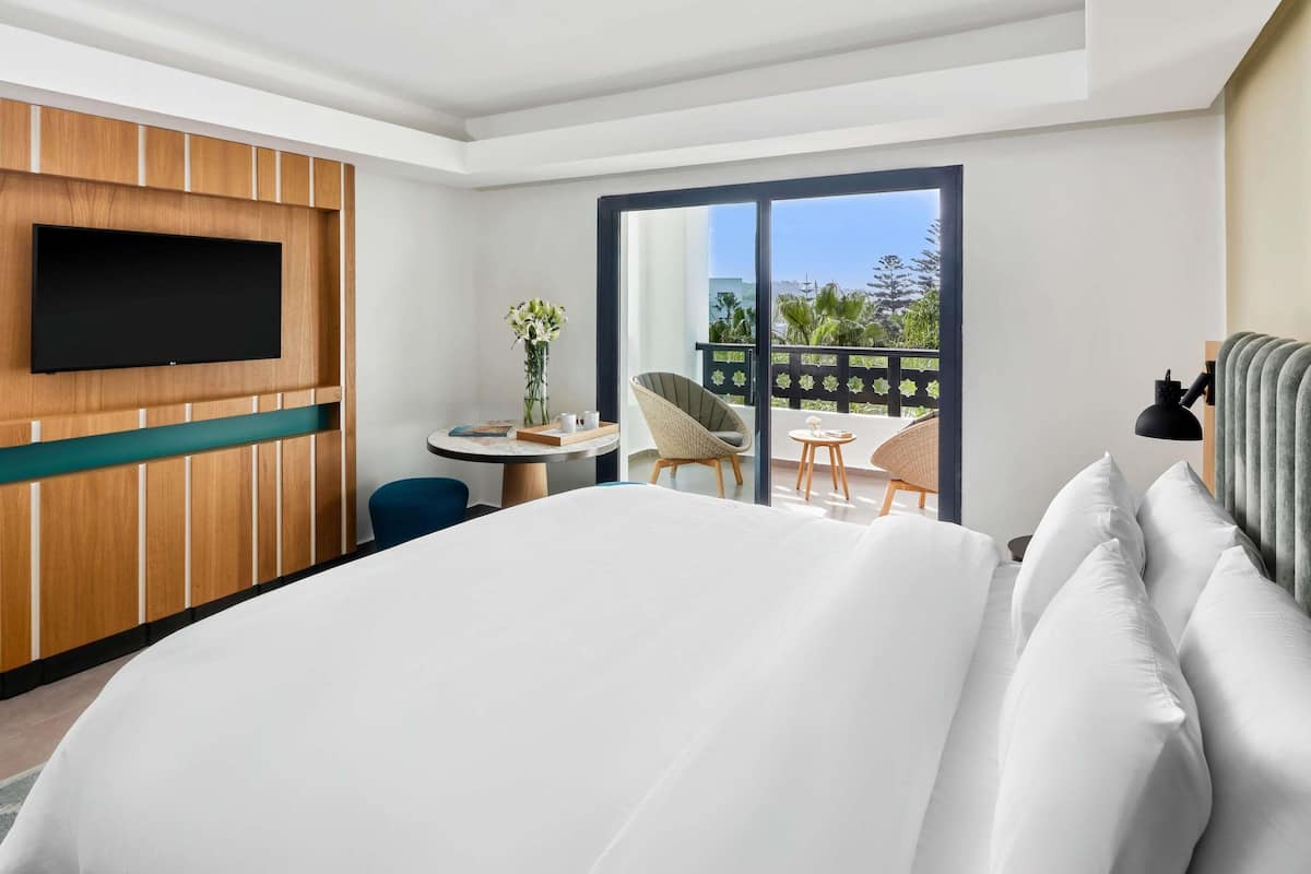 A hotel room with a large bed, a TV mounted on the wall, a small table with a vase of flowers, two wicker chairs, and a balcony with a view.