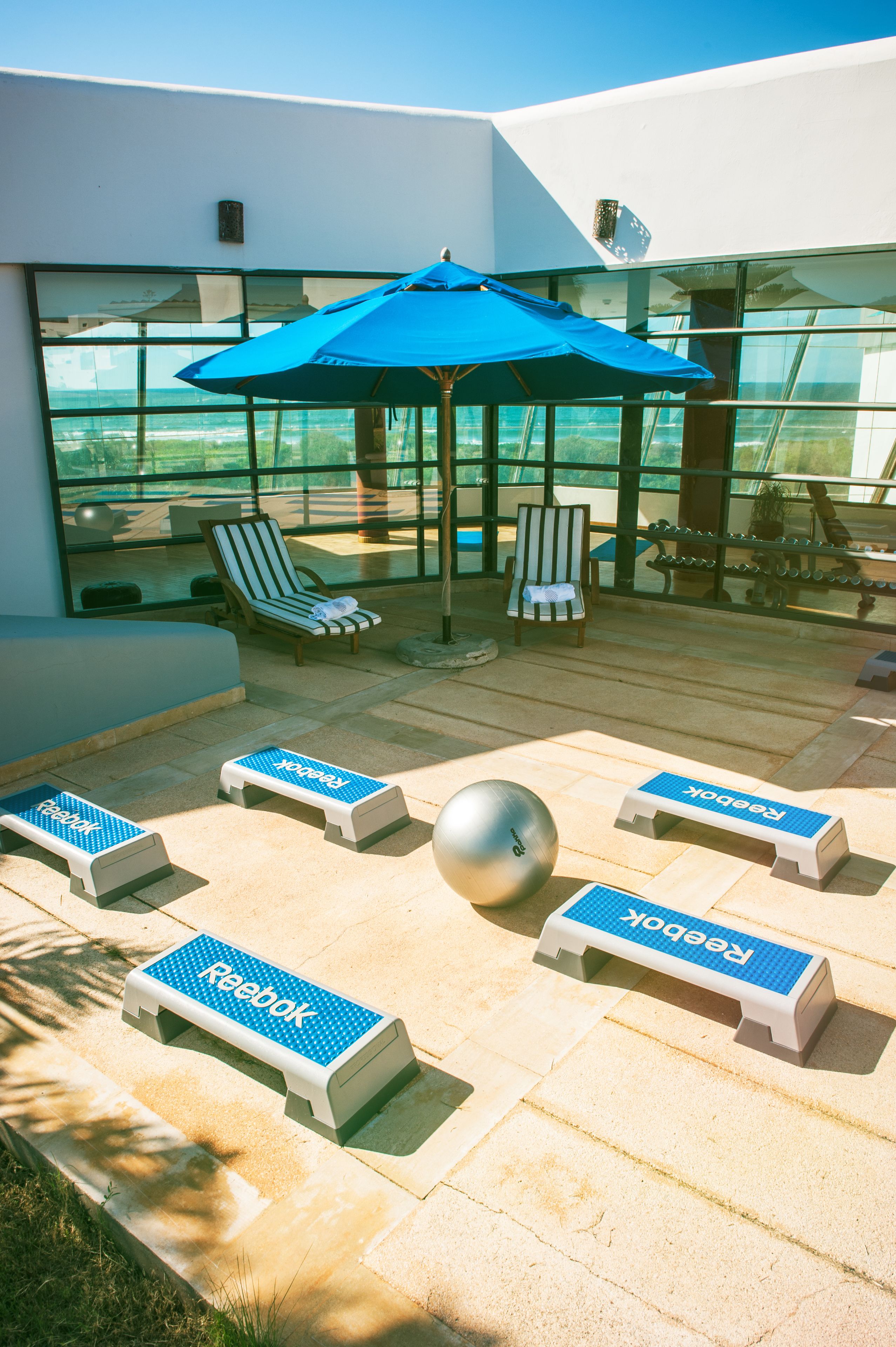 A modern outdoor lounge area with seating, a blue umbrella, and a view through large windows.