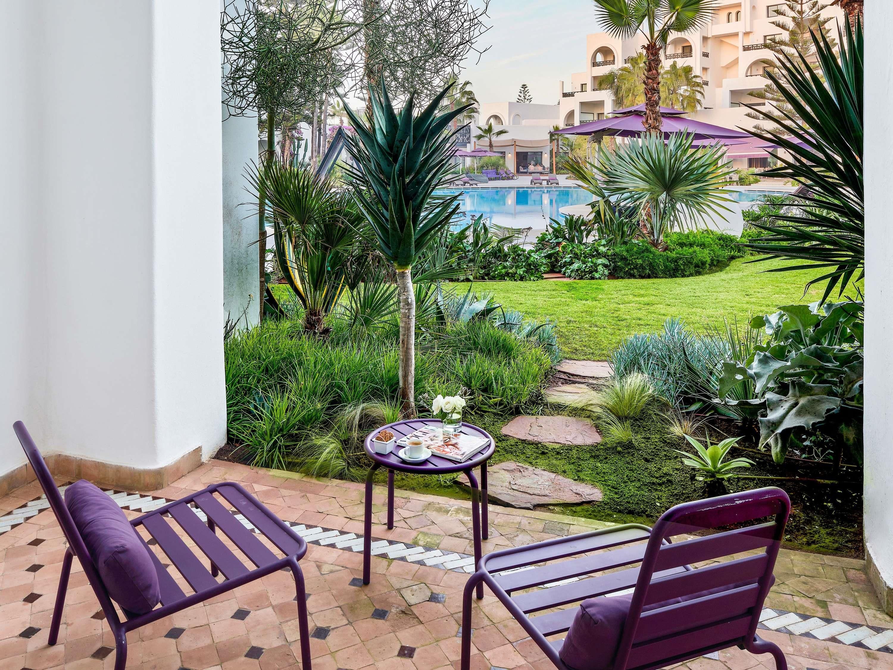 Un patio con dos sillas moradas, una mesa pequeña con una bandeja de comida y vistas a una piscina y follaje.