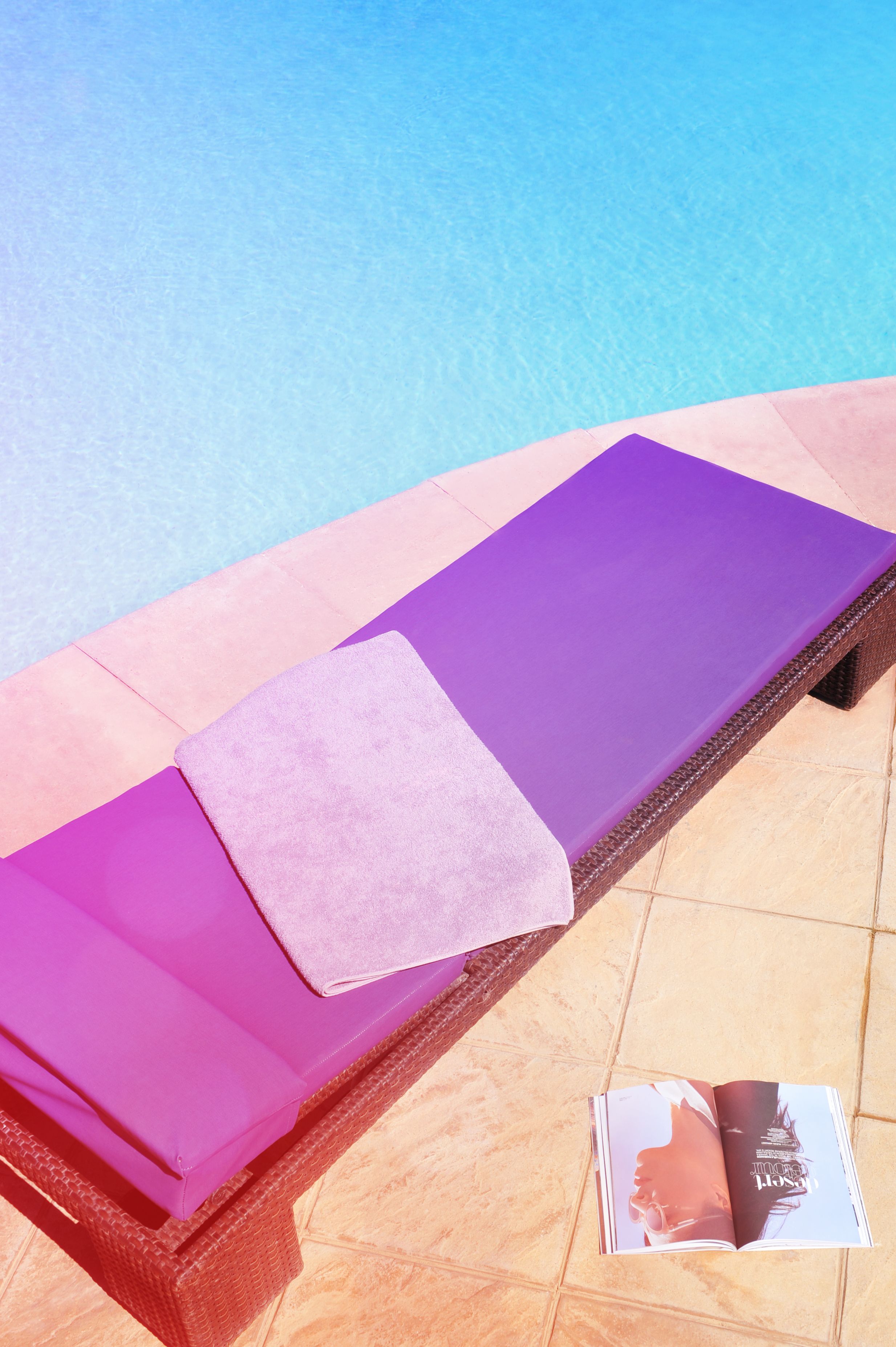 Une chaise longue au bord de la piscine, avec un coussin violet et une serviette rose, et un magazine posé sur le sol carrelé.