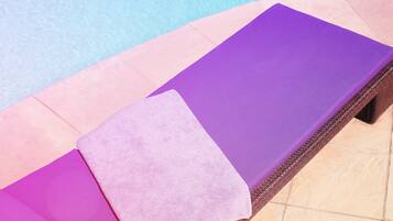 Une chaise longue au bord de la piscine, avec un coussin violet et une serviette rose, et un magazine posé sur le sol carrelé.