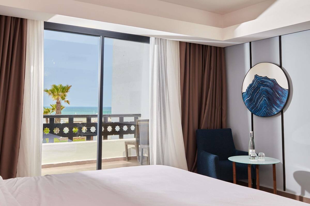 Een hotelkamer met een bed, een stoel, een klein tafeltje met een fles en glazen, een ronde spiegel, en een balkon met uitzicht op zee.