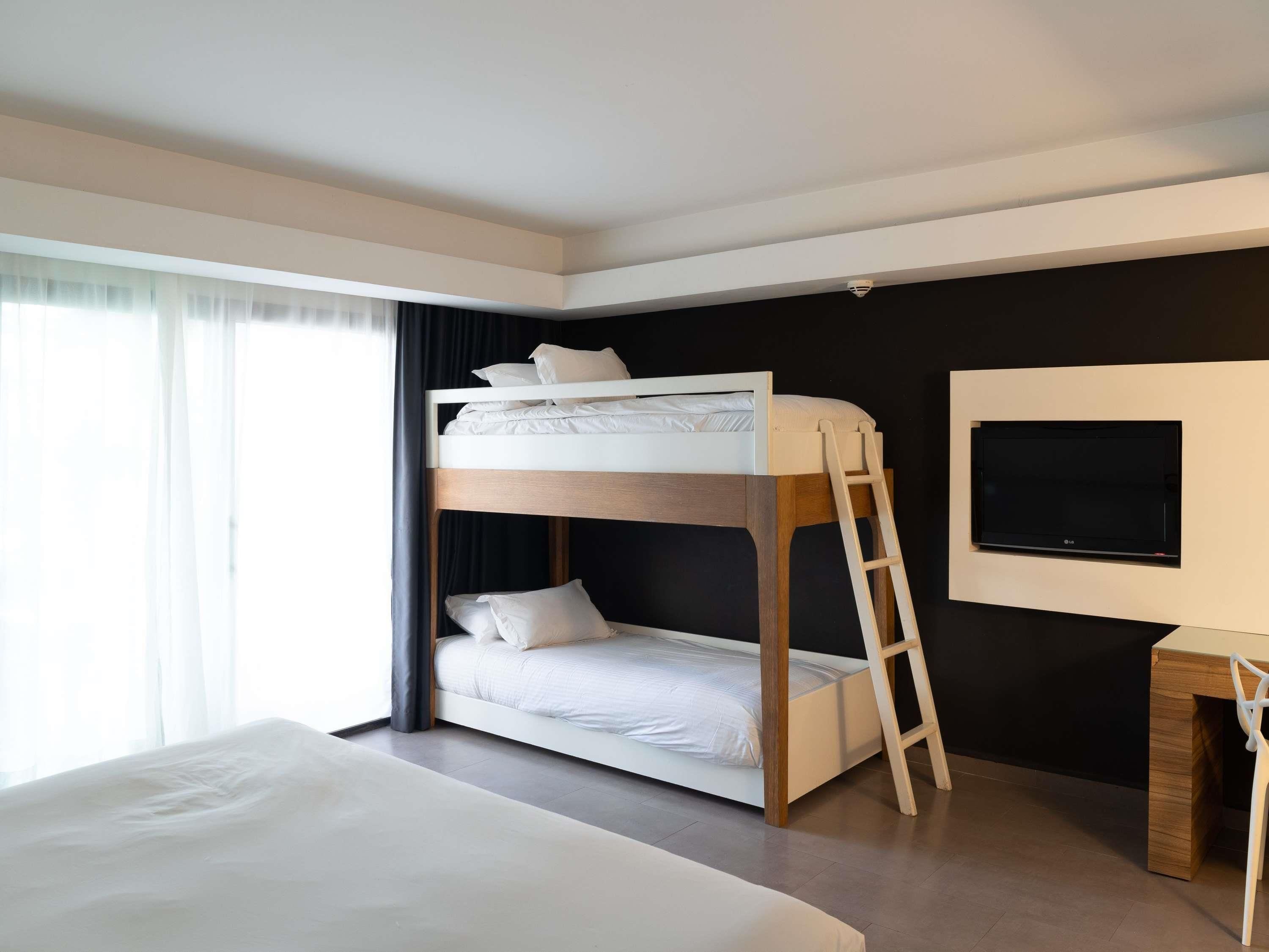 Una cama individual superpuesta con televisión y escritorio, una ventana grande con cortinas y una pared oscura.
