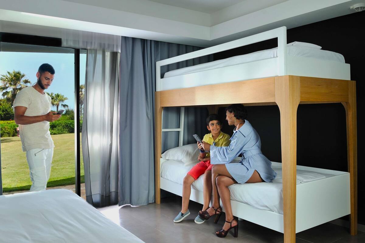 Een slaapkamer met een stapelbed, een groot raam, een bed en een persoon bij het raam.