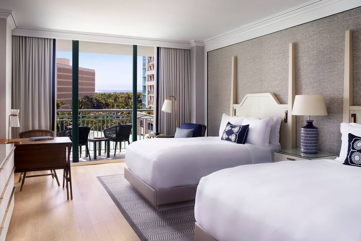 A hotel room with two beds, a desk, and a balcony with a view of buildings and palm trees.