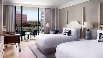 Habitación de hotel con dos camas, un escritorio y un balcón con vistas a edificios y palmeras.